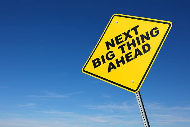 A  yellow road sign indicates that the next big thing is ahead in front of a blue sky with wispy clouds at the horizon. The perspective is from a low camera angle. Road sign is placed on right side of image allowing for copy space on the left side of image.